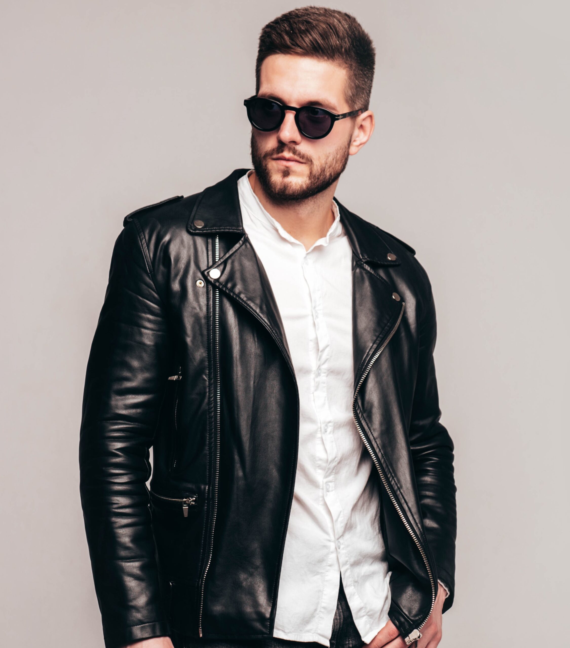 Portrait of handsome confident model. Sexy stylish man dressed in biker leather jacket and black jeans. Fashion hipster male isolated on grey background in studio in sunglasses. Isolated